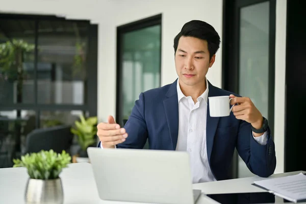 stock image Focused and smart millennial Asian businessman or male CEO in formal business suit sits at this desk, sipping coffee, while having an online meeting with his management team.