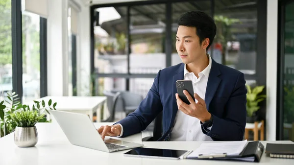 집중적 밀레니엄 아시아 사업가나 관리자는 스마트폰을 사무실에 책상에 컴퓨터를 합니다 — 스톡 사진