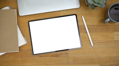 Tahta arka planda dijital tablet beyaz ekran modeli, stilus kalem ve aksesuarları olan bir çalışma alanının üst görünümü.