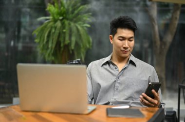Zeki ve yakışıklı Asyalı iş adamı akıllı telefonuyla dışarıdaki bir kafede dizüstü bilgisayarıyla otururken mesajlarını kontrol ediyor. Kentsel yaşam tarzı konsepti