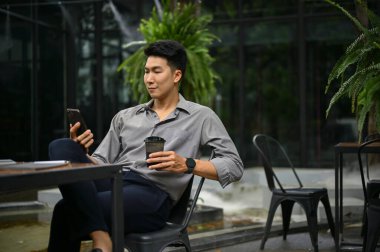 Rahat ve yakışıklı Asyalı adam akıllı telefonunu kullanıyor ve açık havada kahve içiyor..