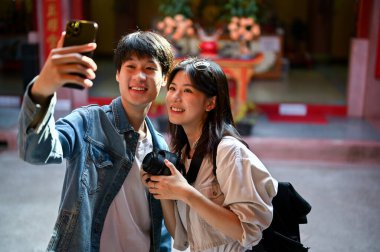 Şirin ve mutlu Asyalı genç turist çift, Çin tapınağını ve eski bir şehri gezerken akıllı telefonlarıyla fotoğraflarını çekiyorlar..