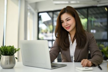 Çekici Asyalı kadın muhasebeci dizüstü bilgisayarını kullanıyor ve ofisinde masasında çalışıyor..