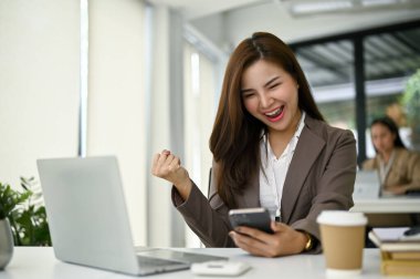 Heyecanlı ve neşeli Asyalı iş kadını ya da ofis çalışanı masasında telefonundaki mesaja bakarken seviniyor ve iyi haberi kutluyor..