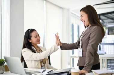 İki neşeli ve mutlu Asyalı iş kadını ofiste çalışırken birbirlerini neşelendirmek için beşlik çakıyor..