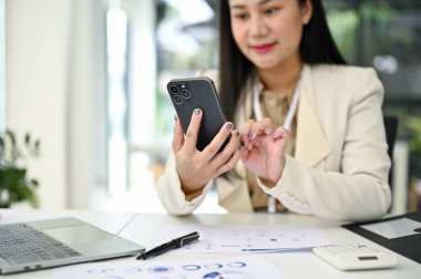 Asyalı profesyonel bir iş kadını ya da kadın muhasebecinin masasındaki akıllı telefonu kullanarak telefonundan birine mesaj gönderdiği yakın plan görüntüsü..