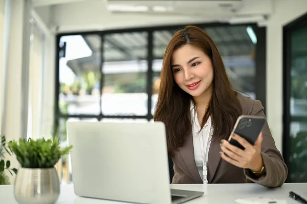 彼女の机のラップトップコンピュータで彼女の仕事をしている間彼女のスマートフォンを保持している魅力的で幸せな千年のアジアの実業家 — ストック写真