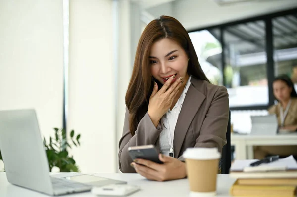 彼女の机に座っている間 彼女の携帯電話に予期せぬメッセージを受信し 興奮し 驚き千年のアジアのビジネスマンの女性 — ストック写真