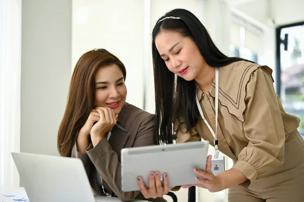 A professional millennial Asian female secretary shows something on her tablet screen to her boss, discussing and planning a new project in the office together.