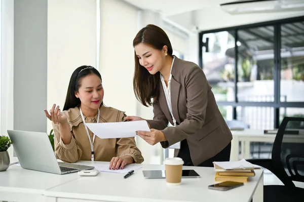 A beautiful young Asian female secretary is showing something on document to her boss, listening to her boss brief her on a new project, and working in the office together.
