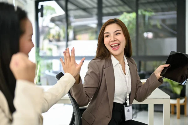 Excited and cheerful young Asian businesswomen giving high fives to each other, celebrating their career success in the office together.