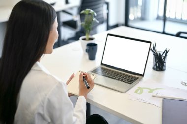 Arka plan görüntüsü, profesyonel ve odaklanmış Asyalı bir iş kadını ya da ofis çalışanı masadaki dizüstü bilgisayarına bakarken not alıyor..