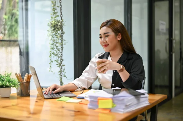 ラップトップコンピュータで仕事をし オフィスの彼女の机でコーヒーを飲みながら彼女のビジネスの仕事に取り組んでいる幸せと魅力的な千年のアジアの実業家 — ストック写真