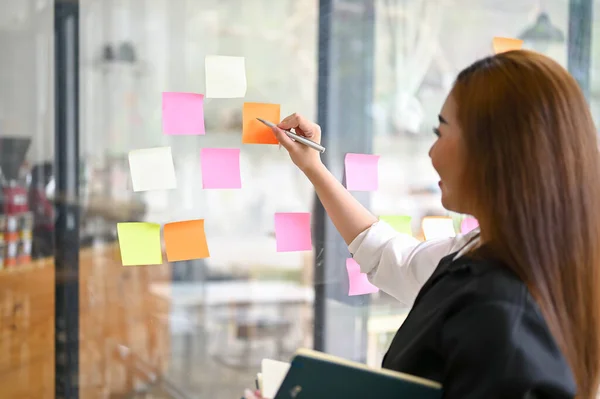 Close-up image of a creative millennial Asian female marketing assistant writing her creative idea on a sticky note on the glass wall.