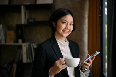 Resmi takım elbiseli başarılı ve kendinden emin Asyalı bir iş kadınının portresi, kameraya gülümsüyor ve elinde bir kahve fincanı ve akıllı telefonuyla modern ofisinde duruyor..