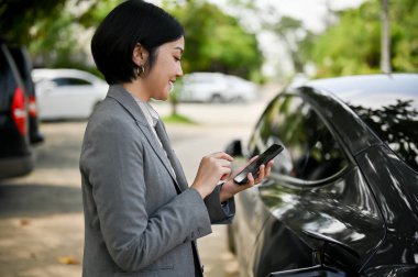 Başarılı bir Asyalı iş kadını, elektrikli arabasının şarj edilmesini beklerken akıllı telefonuyla mesajları kontrol eder. İş insanları ve ulaşım kavramları