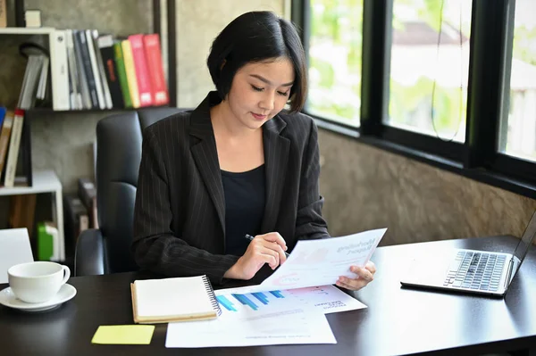 A professional and focused millennial Asian female executive manager is reviewing marketing reports for the new campaign at her desk in her private office.