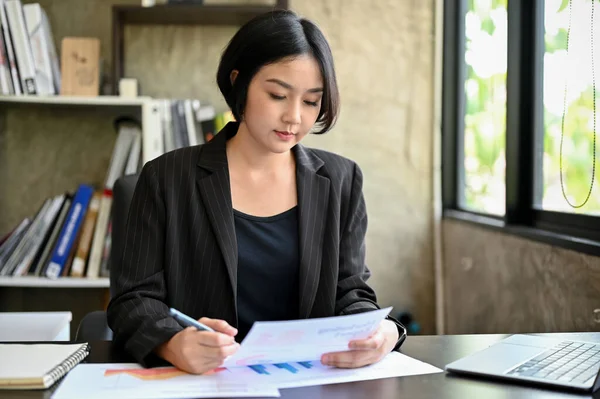 A professional and attractive millennial Asian female executive manager is reviewing marketing reports for the new campaign at her desk in her private office.