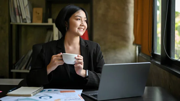 Charming and relaxed Asian businesswoman or female boss in a black suit enjoys her morning coffee at her desk.