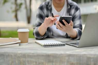 Akıllı telefonunu kullanan, arkadaşlarıyla sohbet eden ya da kampüs parkında otururken internetten bilgi arayan Asyalı bir üniversite öğrencisi görüntüsü..