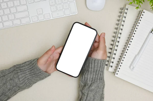 Rahat gri kazaklı bir kadının, ofis malzemeleriyle dolu gri çalışma masasının üzerinde akıllı telefon beyaz ekran maketi tutarken üst görüntüsü..