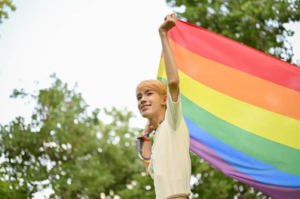 一个笑着 无忧无虑 挂着Lgbt彩虹旗的年轻男同性恋站在绿地里 感到自由和自豪的画像 与Lgbtq Pride一起参加了为期一个月的游行 — 图库照片