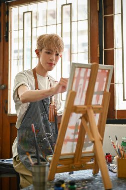 Önlük takan yaratıcı ve çekici Asyalı genç eşcinsel bir adamın portresi stüdyodaki tuval sehpasına resmini çizmeye odaklanıyor..