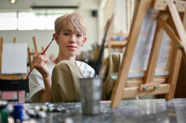 Çekici ve yaratıcı Asyalı genç bir eşcinsel ya da eşcinsel stüdyoda bir sanat atölyesine katılıyor, tuval sehpasının önünde oturuyor..