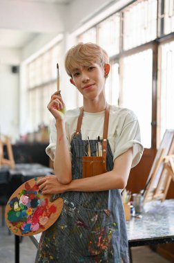 Yetenekli ve yaratıcı eşcinsel bir adamın portresi ya da önlüklü eşcinsel bir sanatçı elinde boya fırçası ve sanat paletiyle yaratıcı stüdyosunda duruyor. LGBTQ + ve yaratıcı iş kavramları