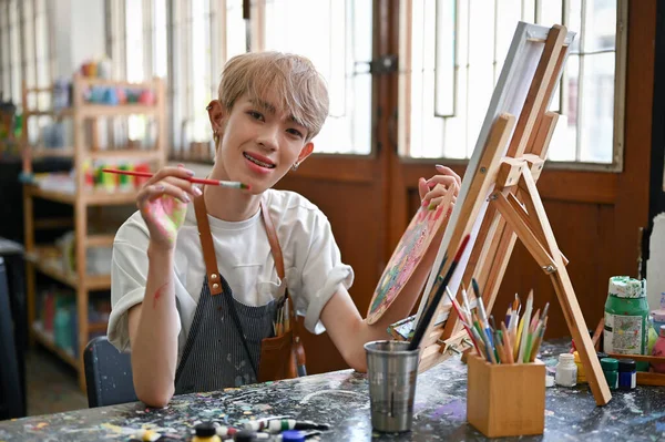 一位快乐而富有创造力的年轻亚洲同性恋艺术学生在工作室的书桌边的一张小画布上画了水彩画 — 图库照片