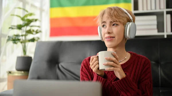 在客厅里通过耳机听音乐的同时 一位悠闲而沉着的年轻的亚洲同性恋男子正在享受他的咖啡 生活方式概念 — 图库照片