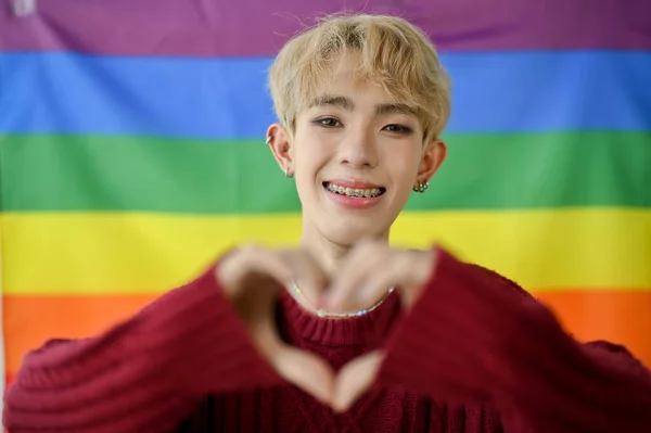 一位有魅力 面带微笑的年轻亚洲同性恋者或同性恋男子站在房间里时 在墙上挂着一面Lgbt彩虹色的旗帜 并展示了他的心迹 — 图库照片