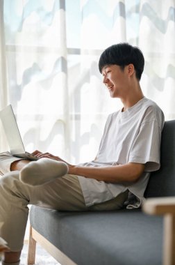 Yandan bakıldığında, sıradan giyinmiş mutlu bir Asyalı, oturma odasındaki bir kanepede dinlenirken sosyal medyada sosyal medyada videolar izliyor..