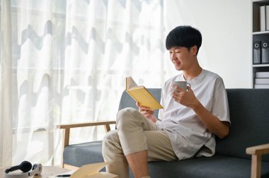 Günlük kıyafetlerini giymiş mutlu bir Asyalı sabah kahvesini yudumluyor ve oturma odasındaki kanepede kitap okuyor. Yaşam tarzı kavramı