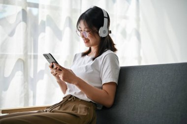 Gündelik giysiler içinde güzel bir Asyalı kadının arka görüntüsü, oturma odasındaki bir kanepede arkadaşlarıyla akıllı telefonuyla sohbet ederken kulaklıkla müzik dinlemekten zevk alıyor..