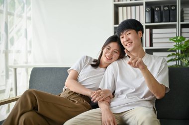 Genç ve tatlı Asyalı bir kız arkadaş, erkek arkadaşına sarılıyor ve birlikte oturma odasında rahat bir kanepede televizyon izliyorlar..