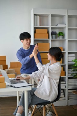 Neşeli ve mutlu bir Asyalı genç adam, çevrimiçi bir dükkan sahibi, kız arkadaşına beşlik çakıyor, ev ofislerinde birlikte çalışırken online iş başarılarını kutluyor..