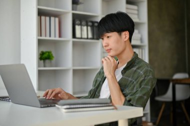 Zeki ve profesyonel bir Asyalı erkek ofis çalışanı veya asistanı modern bir ofiste masasında oturur ve dizüstü bilgisayarına odaklanır..