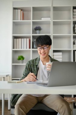 Zeki ve yakışıklı Asyalı genç bir ofis çalışanının portresi ya da pazarlama asistanı masasında çalışıyor, kahvesini yudumlarken not defterindeki detayları kontrol ediyor..
