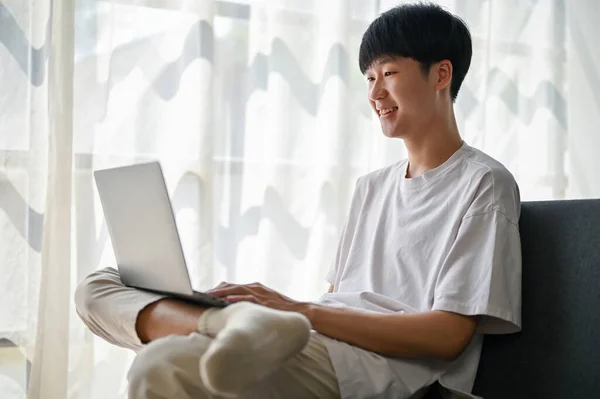 一个快乐而英俊的亚洲年轻人在客厅的沙发上用手提电脑 这让人回想起他 — 图库照片