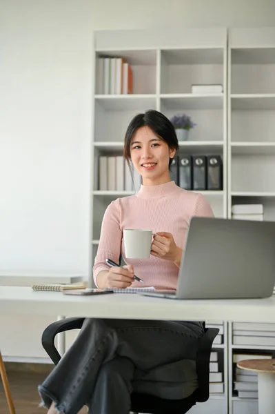 美しいと成功した若いアジアのビジネスマンの肖像画カジュアルな服で彼女の机の上に座っている彼女の手にコーヒーカップ — ストック写真