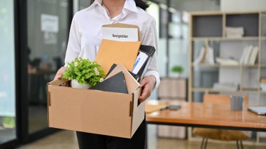 Ofiste elinde bir karton kutu ve istifa mektubuyla duran bir kadın ofis çalışanının kesilmiş görüntüsü. İstifa et, işi bırak, kovuldun, işsizsin.