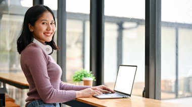 Gayri resmi giysili güzel bir Asyalı kadının arka görüntüsü dizüstü bilgisayarıyla pencerenin karşısında oturuyor..