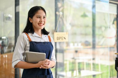 Önlüklü güzel ve arkadaş canlısı Asyalı bir bayan garson elinde dijital tabletle kahve dükkanının girişinde duruyor..