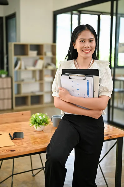 Attractive and successful Asian businesswoman or female office worker stands in her office with document folders in her hands.