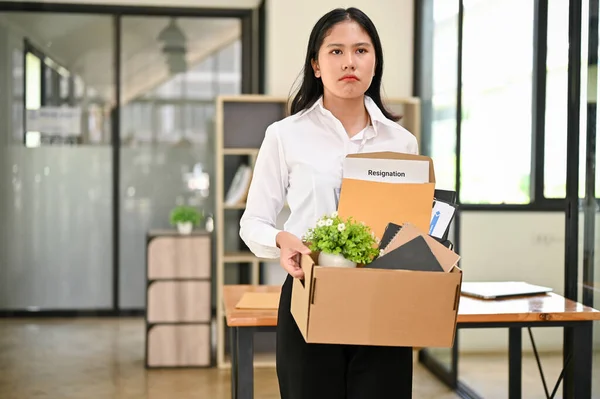 Sad and upset young Asian female office worker stands in the office with a cardboard box with her stuff and her resignation letter. resign, leave the job, fired, unemployed