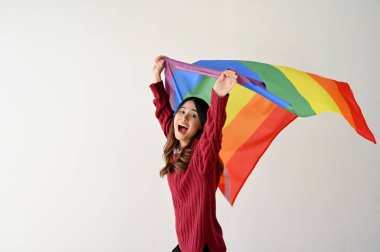 Gökkuşağı renkli LGBT bayrağı taşıyan neşeli ve neşeli Asyalı bir kadın izole edilmiş beyaz bir stüdyo arka planının üzerinde duruyor. LGBTQ + gurur ayı