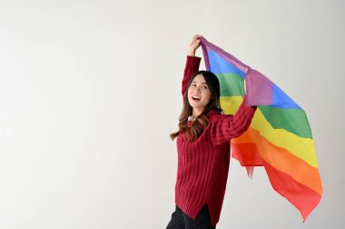 Gökkuşağı renkli LGBT bayrağı taşıyan sıradan giyinmiş mutlu ve güzel bir Asyalı kadın izole edilmiş beyaz bir stüdyo arkasında duruyor. LGBTQ + gurur ayı