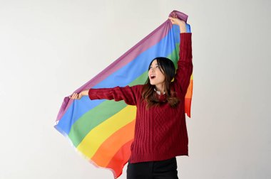 Gökkuşağı renkli LGBT bayrağı taşıyan sıradan giyinmiş mutlu bir Asyalı kadın izole edilmiş beyaz bir stüdyo arkasında duruyor. LGBTQ + gurur ayı, özgürlük, insan hakları