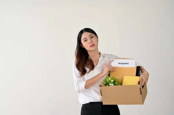 stock image A tired and despairing young Asian businesswoman or female office worker holds a cardboard box with her stuff and a resignation letter, standing over an isolated white background.